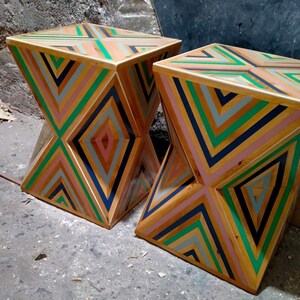 K&ouml;nnte beinhalten: Zwei Holzhocker mit einem geometrischen Muster aus rosa, gr&uuml;nen, blauen und grauen Streifen. Die Hocker sind aus recyceltem Holz gefertigt und haben ein einzigartiges, modernes Design.
