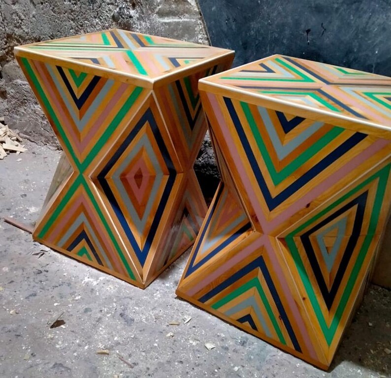 K&ouml;nnte beinhalten: Zwei Holzhocker mit einem geometrischen Muster aus rosa, gr&uuml;nen, blauen und grauen Streifen. Die Hocker sind aus recyceltem Holz gefertigt und haben ein einzigartiges, modernes Design.