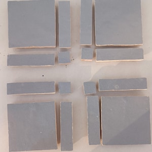 May include: Arrangement of gray ceramic tiles in various shapes and sizes. The tiles include squares, rectangles, and smaller square pieces, all with a matte finish and visible edges. The tiles are arranged on a neutral surface.