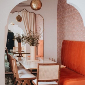 May include: A dining area with a white marble table and wooden chairs. The chairs have woven cane seats. A large, orange-red banquette is on the right side of the image. The room has a green tiled floor and a white arched doorway.