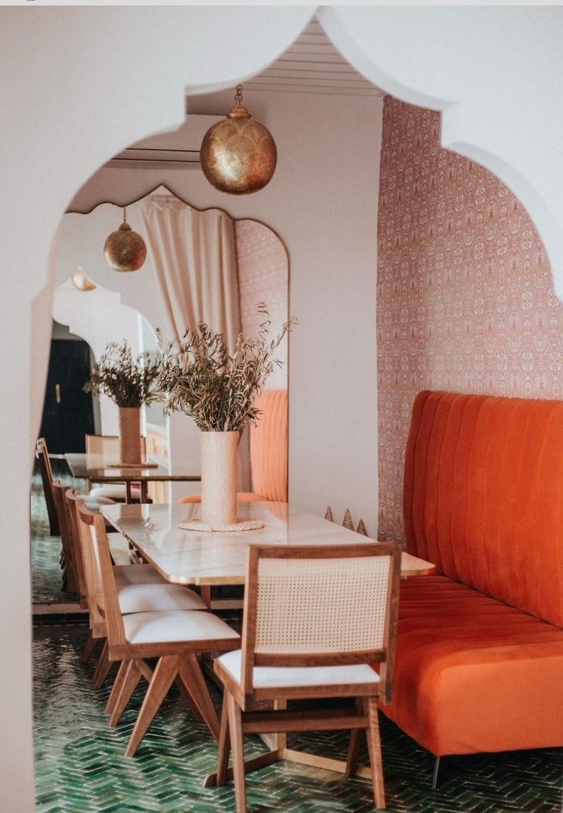 May include: A dining area with a white marble table and wooden chairs. The chairs have woven cane seats. A large, orange-red banquette is on the right side of the image. The room has a green tiled floor and a white arched doorway.