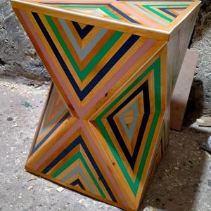 K&ouml;nnte beinhalten: Ein geometrischer Holzhocker mit einem einzigartigen Design mit abwechselnden Streifen aus rosa, gr&uuml;nem, blauem und grauem Holz. Der Hocker hat eine quadratische Oberseite und eine leicht sanduhrf&ouml;rmige Form.