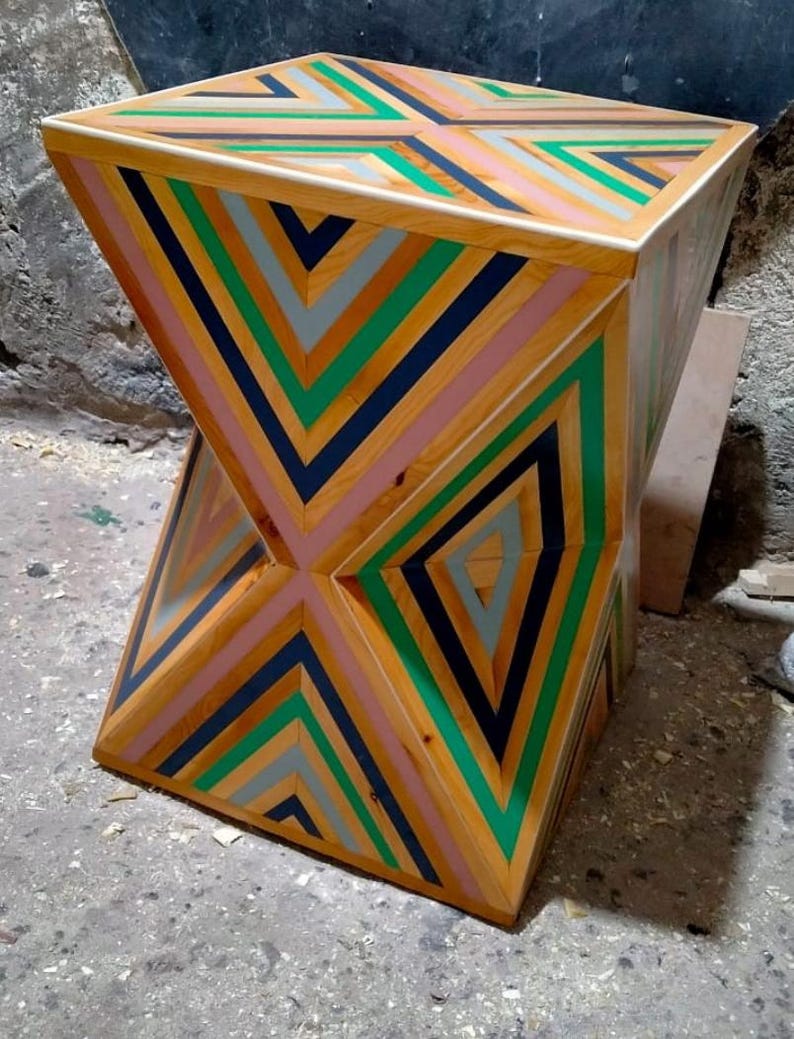 K&ouml;nnte beinhalten: Ein geometrischer Holzhocker mit einem einzigartigen Design mit abwechselnden Streifen aus rosa, gr&uuml;nem, blauem und grauem Holz. Der Hocker hat eine quadratische Oberseite und eine leicht sanduhrf&ouml;rmige Form.