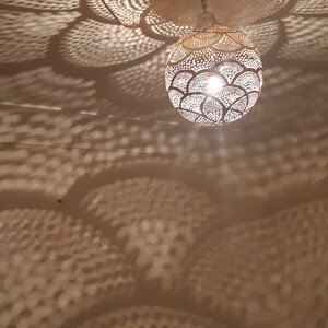 May include: A white, round, metal pendant light fixture with a decorative, cut-out design casts a patterned shadow on the ceiling. The light fixture is illuminated, creating a warm glow.