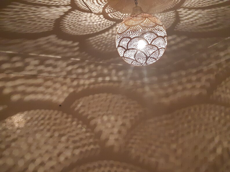 May include: A white, round, metal pendant light fixture with a decorative, cut-out design casts a patterned shadow on the ceiling. The light fixture is illuminated, creating a warm glow.