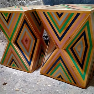 K&ouml;nnte beinhalten: Zwei Holzhocker mit einem geometrischen Muster aus gr&uuml;nen, blauen, rosa und grauen Streifen. Die Hocker sind aus Holz gefertigt und haben ein einzigartiges, modernes Design.