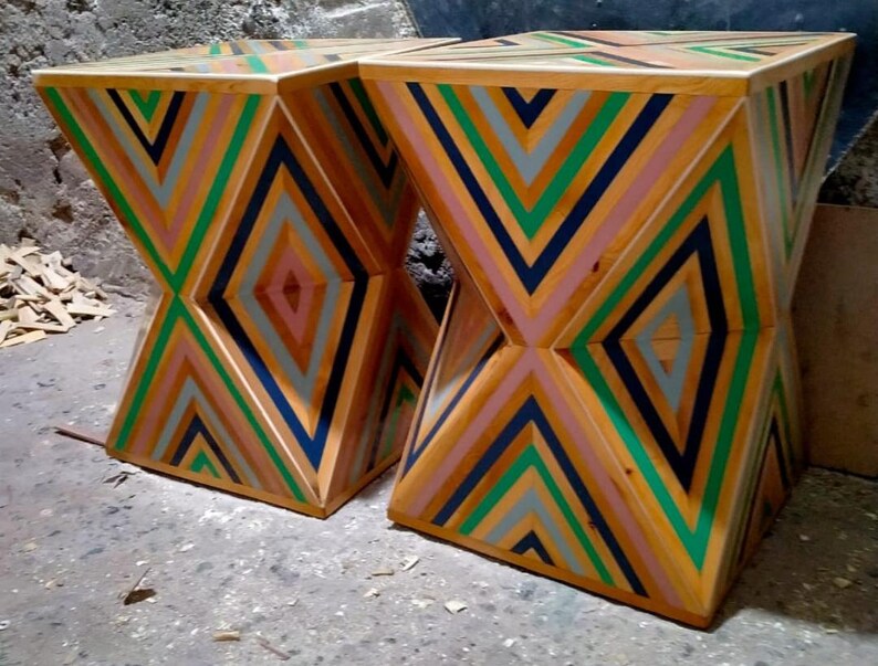 K&ouml;nnte beinhalten: Zwei Holzhocker mit einem geometrischen Muster aus gr&uuml;nen, blauen, rosa und grauen Streifen. Die Hocker sind aus Holz gefertigt und haben ein einzigartiges, modernes Design.
