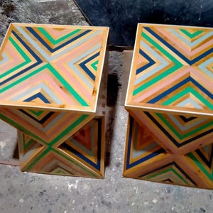 K&ouml;nnte beinhalten: Zwei geometrische Beistelltische aus Holz mit einem farbenfrohen, gestreiften Design. Die Tische sind aus Holz und haben ein geometrisches Muster aus rosa, gr&uuml;nen, blauen und wei&szlig;en Streifen.