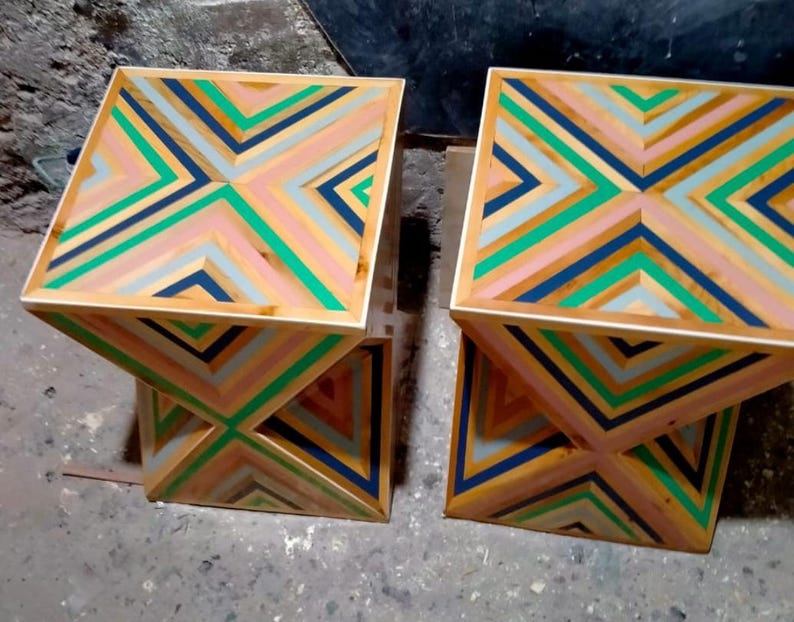 K&ouml;nnte beinhalten: Zwei geometrische Beistelltische aus Holz mit einem farbenfrohen, gestreiften Design. Die Tische sind aus Holz und haben ein geometrisches Muster aus rosa, gr&uuml;nen, blauen und wei&szlig;en Streifen.