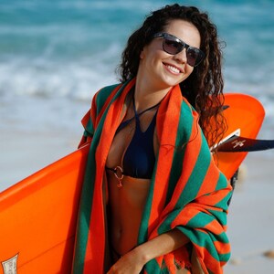 Puede incluir: Una mujer con gafas de sol y un pareo a rayas verdes y naranjas sostiene una tabla de surf naranja brillante.