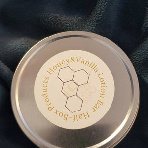 Könnte beinhalten: Eine runde, silberfarbene Dose mit einem cremefarbenen Etikett. Das Etikett zeigt den Text "Honey & Vanilla Lotion Bar" in einem kreisförmigen Design, zusammen mit einer Waben-Grafik und einer Bienen-Illustration. Die Dose liegt auf einer dunkelblauen, strukturierten Oberfläche.