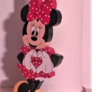 May include: A wooden figurine of Minnie Mouse in a red dress with white polka dots. She is holding her hands in a heart shape and has a red heart on her dress. The figurine is standing on a wooden base.