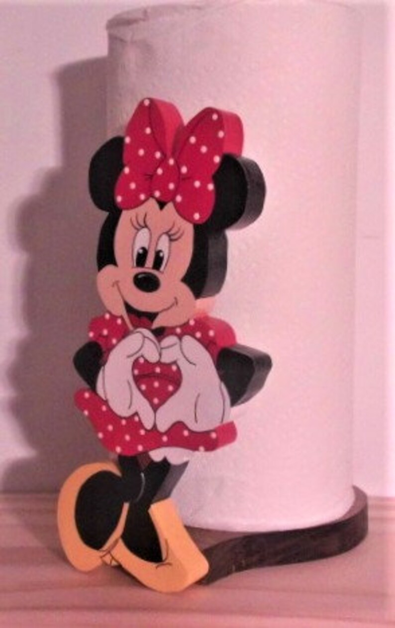 May include: A wooden figurine of Minnie Mouse in a red dress with white polka dots. She is holding her hands in a heart shape and has a red heart on her dress. The figurine is standing on a wooden base.