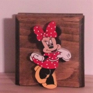 May include: A wooden wall decor featuring a 3D cutout of Minnie Mouse. Minnie is wearing a red dress with white polka dots and a red bow with white polka dots. She has black shoes with yellow soles.
