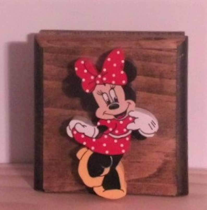 May include: A wooden wall decor featuring a 3D cutout of Minnie Mouse. Minnie is wearing a red dress with white polka dots and a red bow with white polka dots. She has black shoes with yellow soles.