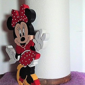 Puede incluir: Un porta rollo de papel de cocina de madera con un personaje de dibujos animados rojo, blanco y negro con un lazo rojo y lunares blancos. El personaje sostiene un rollo de papel de cocina blanco.