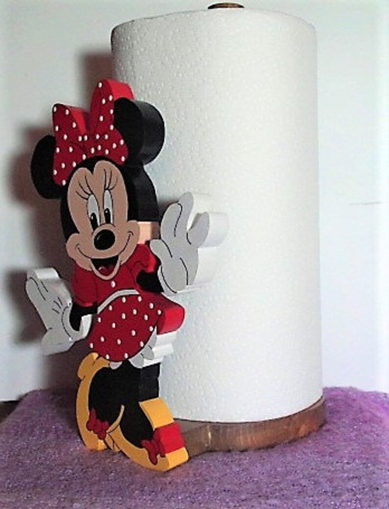 Puede incluir: Un porta rollo de papel de cocina de madera con un personaje de dibujos animados rojo, blanco y negro con un lazo rojo y lunares blancos. El personaje sostiene un rollo de papel de cocina blanco.