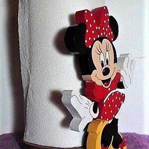 Puede incluir: Un soporte de papel de cocina de madera con una imagen de dibujos animados de Minnie Mouse. Minnie lleva un vestido rojo con lunares blancos y un lazo rojo con lunares blancos. Lleva zapatos negros y tiene una cara blanca y negra.