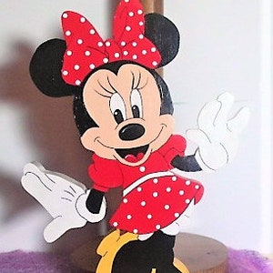 Puede incluir: Un recorte de madera de Minnie Mouse, pintado en rojo, negro y blanco. Lleva un vestido rojo con lunares blancos y un lazo rojo con lunares blancos. Est&aacute; sonriendo y saludando.