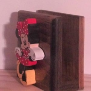May include: A wooden napkin holder with a hand-painted Minnie Mouse design. The holder is made of dark brown wood and features a three-dimensional Minnie Mouse figure with red polka dots, a yellow bow, and white gloves.