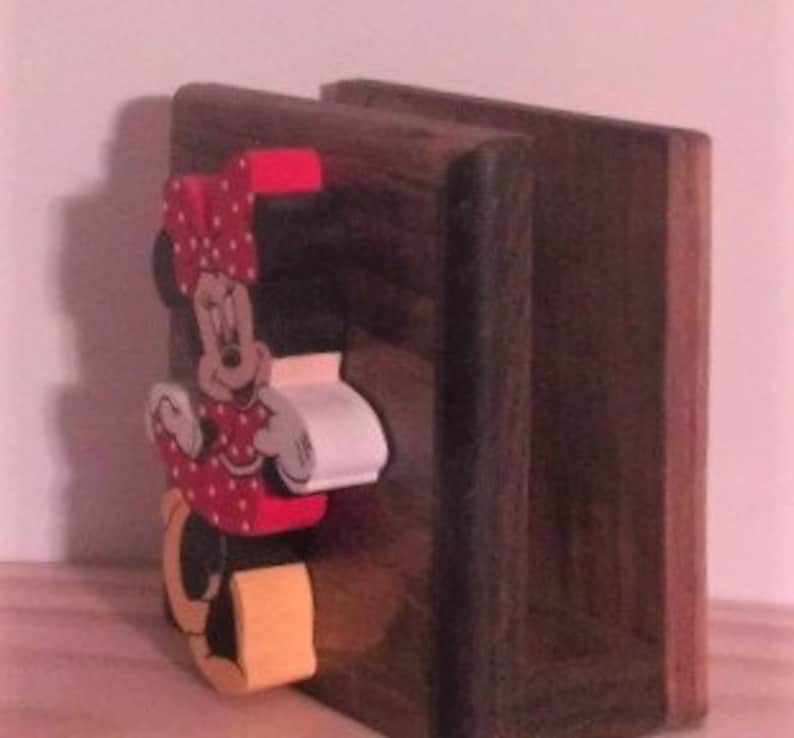 May include: A wooden napkin holder with a hand-painted Minnie Mouse design. The holder is made of dark brown wood and features a three-dimensional Minnie Mouse figure with red polka dots, a yellow bow, and white gloves.