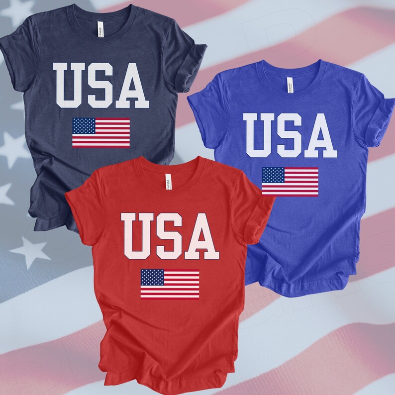 May include: Three t-shirts with the word "USA" printed in white letters and a small American flag below. The shirts are navy blue, royal blue, and red.
