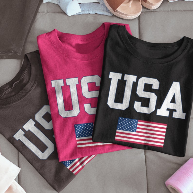 May include: Three t-shirts with the word "USA" printed on them in white letters. The shirts are in brown, pink, and black. Each shirt has a small American flag printed on it.
