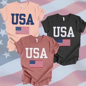 May include: Three t-shirts with the word "USA" printed on them in different colors. The shirts are all in a casual style and feature a small American flag printed below the word "USA".