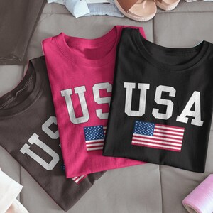 May include: Three t-shirts with the word "USA" printed on them in white letters. The shirts are in different colors: pink, black, and brown. Each shirt has a small American flag printed on it.