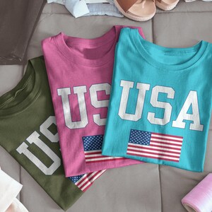 May include: Three t-shirts with the word "USA" printed on them in white letters. The shirts are in different colors: pink, blue, and green. Each shirt has a small American flag printed on it.