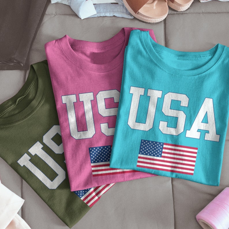 May include: Three t-shirts with the word "USA" printed on them in white letters. The shirts are in different colors: pink, blue, and green. Each shirt has a small American flag printed on it.