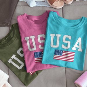 May include: Three t-shirts with the word "USA" printed on them in white letters. The shirts are in different colors: green, pink, and blue. Each shirt has a small American flag printed on it.