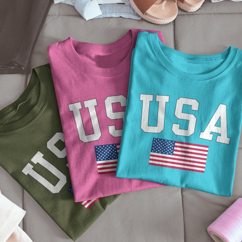 May include: Three t-shirts with the word "USA" printed on them in white letters. The shirts are in different colors: green, pink, and blue. Each shirt has a small American flag printed on it.