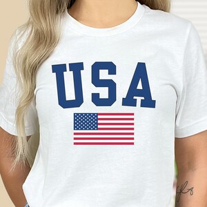 May include: White t-shirt with a blue "USA" graphic and a red, white, and blue American flag.