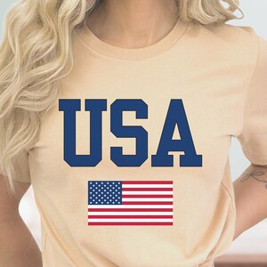 May include: A beige t-shirt with the word "USA" printed in navy blue letters. Below the word is a red, white, and blue American flag.
