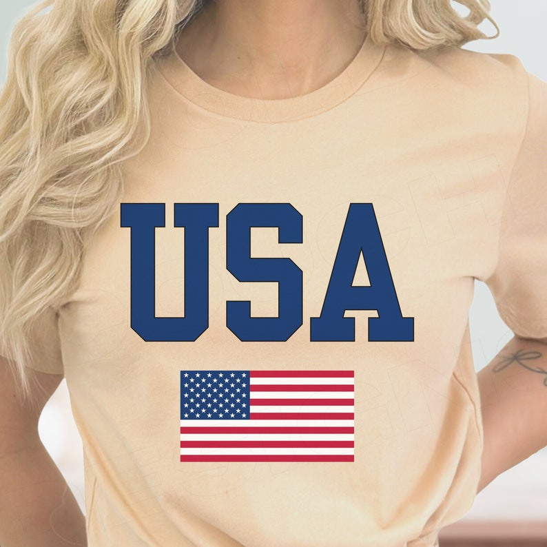 May include: A beige t-shirt with the word "USA" printed in navy blue letters. Below the word is a red, white, and blue American flag.