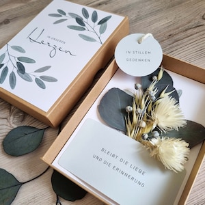 Könnte beinhalten: Eine Trauerkarte mit weißem Hintergrund und braunem Karton. Die Karte zeigt ein Aquarelldesign aus Eukalyptusblättern und dem Text "In unseren Herzen". Der Karton enthält außerdem eine weiße Karte mit dem Text "Bleibt die Liebe und die Erinnerung" und einen kleinen weißen Kreis mit dem Text "In stillem Gedenken".