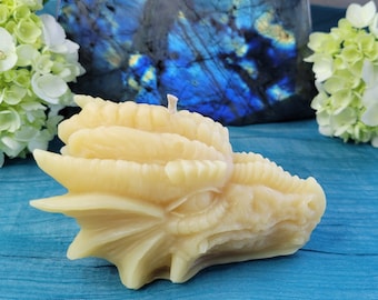 May include: A detailed dragon head candle made of beeswax. The candle is a light yellow colour and has a textured surface. The candle is sitting on a blue surface.