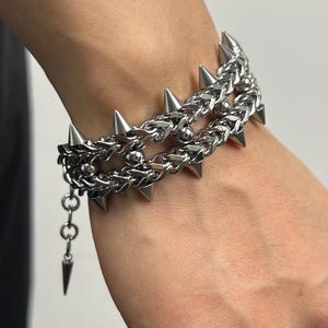 May include: A silver-colored bracelet with a punk rock aesthetic. The bracelet features multiple rows of chain links and is adorned with sharp, conical spikes. The design includes a dangling spike charm, adding to its edgy appeal.