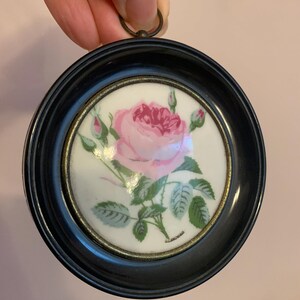 Op de afbeelding: Een ingelijst porseleinen plaatje van een roze roos met groene bladeren en knoppen. De lijst is zwart met een gouden rand.