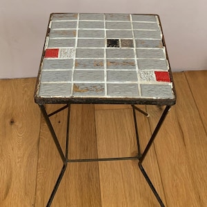 May include: A small square side table with a tiled top. The tiles are mostly gray with some red, black, and white accents. The table has a simple black metal frame with four legs.