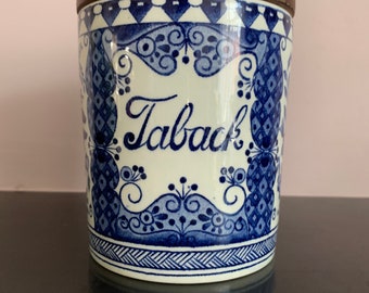 Peut inclure: Un pot à tabac en céramique blanche avec un motif floral bleu et blanc et le mot "Taback" écrit en cursive. Le pot a un couvercle en bois.
