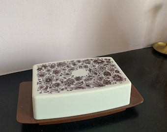 Könnte beinhalten: Eine weiße Keramik-Butterdose mit einem braunen Blumendesign auf dem Deckel. Die Dose steht auf einem braunen Tablett.
