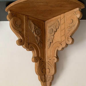 Könnte beinhalten: Ein Eckregal aus Holz mit aufwendigen Schnitzereien. Das Regal ist aus hellbraunem Holz und hat eine abgerundete Kante. Die Schnitzereien sind in Form von Blumen und Blättern.