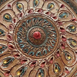 Puede incluir: Un plato circular de terracota con un diseño detallado e intrincado. El diseño presenta un círculo central con un punto rojo, rodeado de círculos concéntricos azules, blancos y dorados. El borde exterior del plato está decorado con un patrón repetitivo de rojo, azul y dorado.