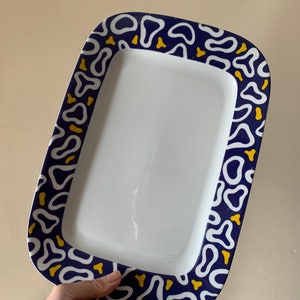 May include: A white rectangular serving platter with a blue and yellow border. The border features a repeating pattern of abstract shapes.