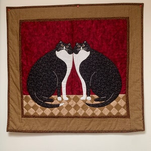 Peut inclure: Une tenture murale matelassée représentant deux chats noirs et blancs avec des accents rouges, bruns et beiges. Les chats se font face et ont une tache blanche sur la poitrine.