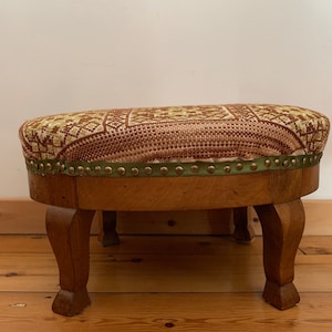 Puede incluir: Un puf redondo con estampado marrón y verde, con estructura de madera y cuatro patas. El puf tiene un ribete de tela verde con tachuelas doradas.