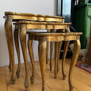 May include: Set of three antique nesting tables. Each table has a scalloped edge and curved legs. The tables are finished in gold with distressed details, adding a vintage aesthetic. The largest table is approximately 24 inches wide.