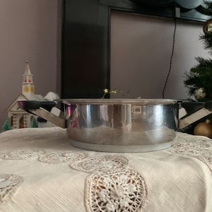 Op de afbeelding: Een stalen pan met zwarte handvatten staat op een witte kanttafelkleed. De pan heeft een glanzend, reflecterend oppervlak.